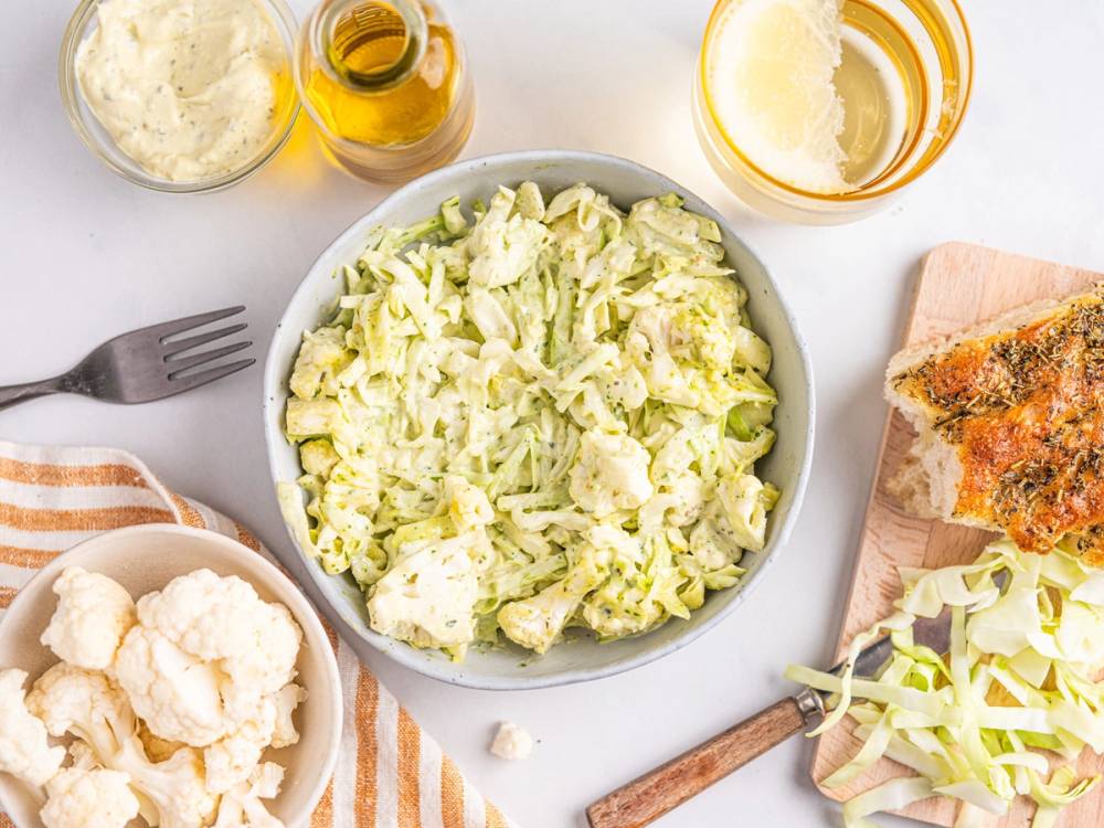 En bolle med fersk, grønn salat omgitt av ulike salatingredienser og dressinger på et kjøkkenbord.