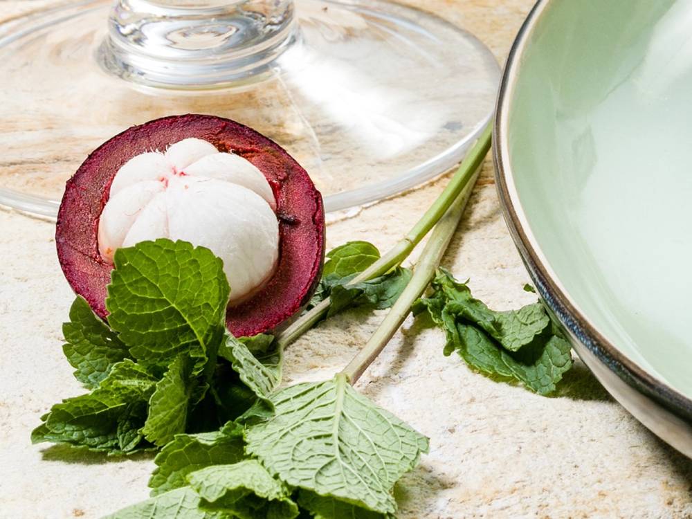 En hel mangostanfrukt med hvit frukt inni, omgitt av grønne mynteblader og en skje, på et bord.