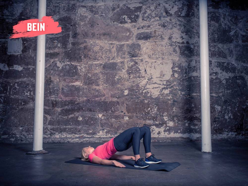 En person ligger på ryggen med knærne bøyd og føttene i gulvet, utfører en setehev-øvelse på en treningsmatte mot en murvegg.