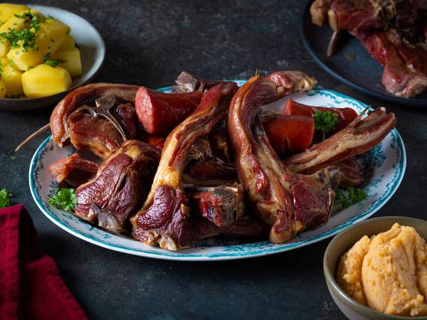 Pinnekjøtt med kålrotstappe på et serveringsfat, omgitt av poteter og annet tilbehør.