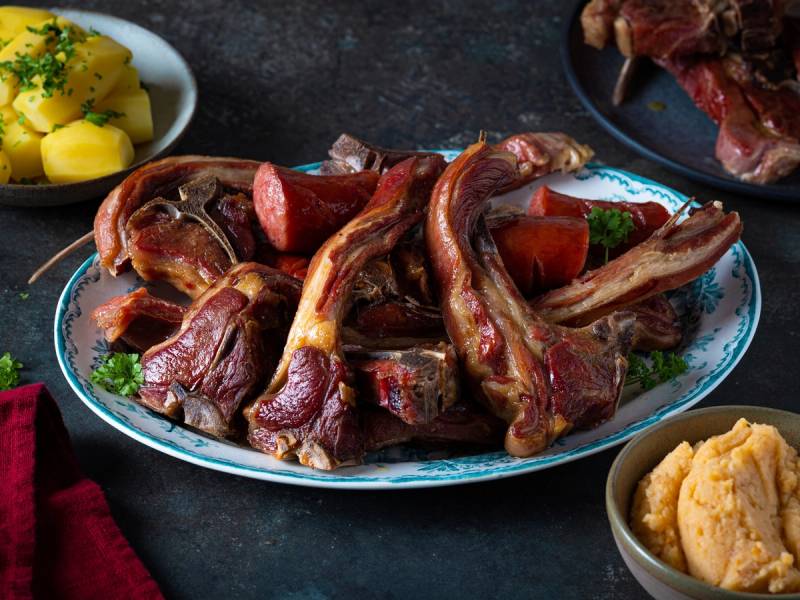 Pinnekjøtt med kålrotstappe på et serveringsfat, omgitt av poteter og annet tilbehør.