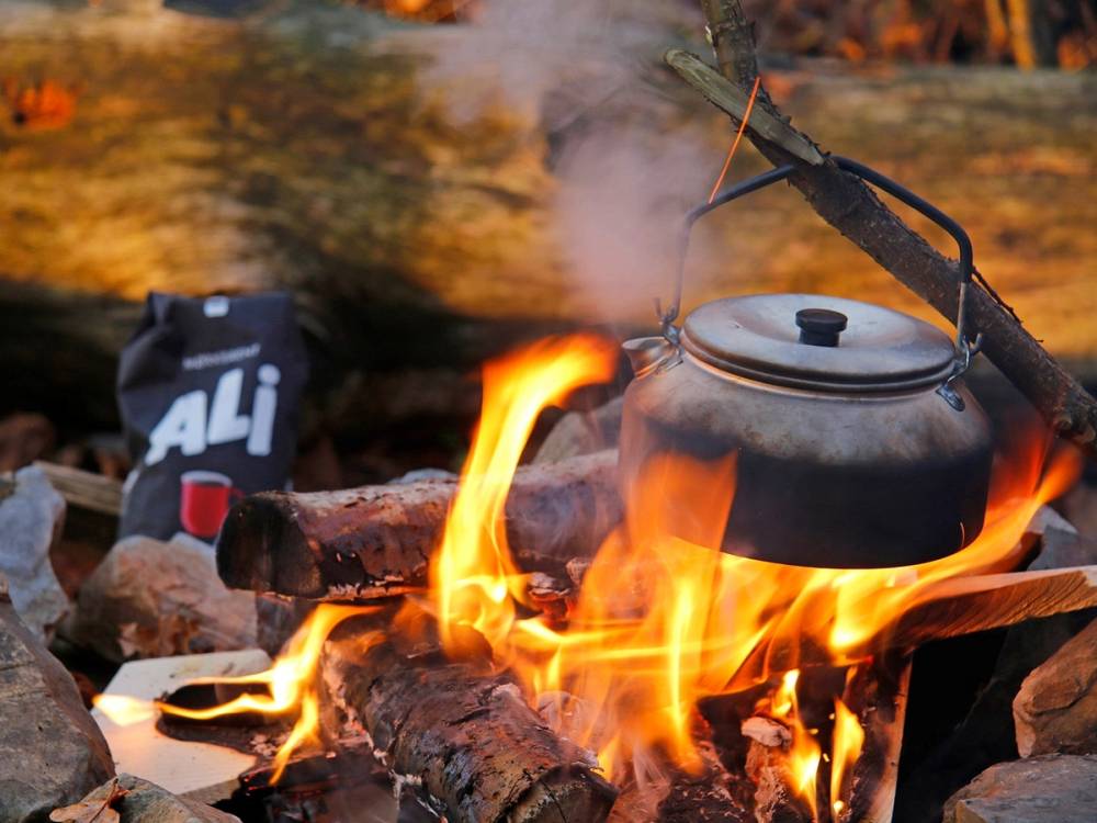 En svart kjele med lokk står på et bål med flammer og røykskyer stiger opp, omgitt av steiner og vedkubber.