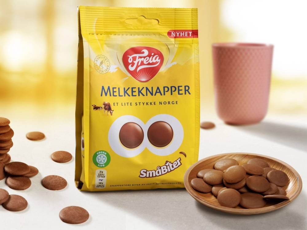 En pose med Freia Melkeknapper ligger på et bord med noen knapper spredt rundt, og en rosa kopp i bakgrunnen.