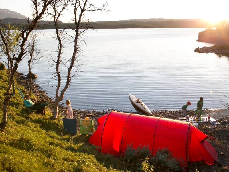 En gruppe mennesker har satt opp et telt ved vannkanten under en solnedgang, med turmat og utstyr rundt seg.