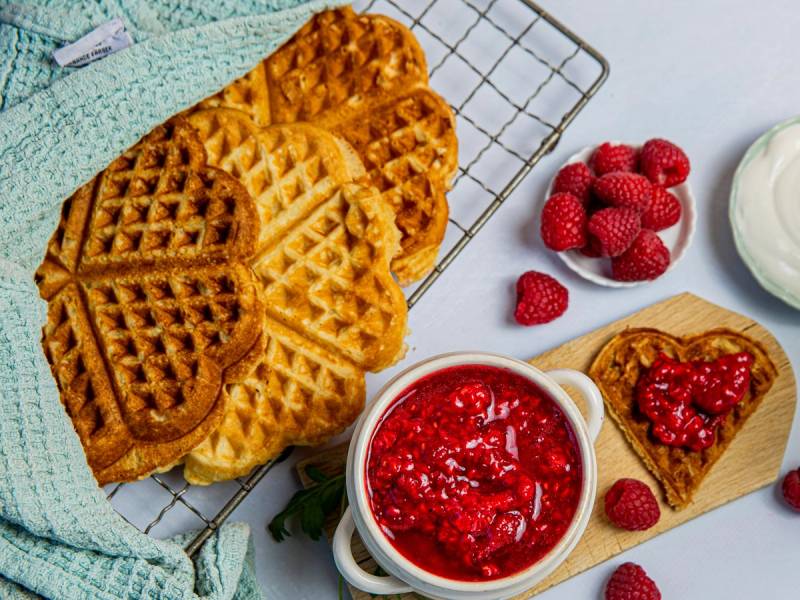 Vafler med bringebærsyltetøy og friske bringebær på et fat, med en skål med bringebærsyltetøy og en liten skål med rømme ved siden av.