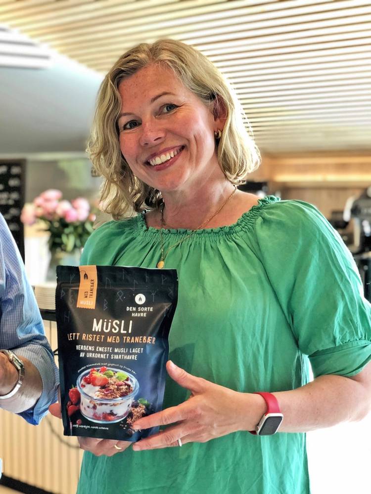 En kvinne i grønn bluse holder en pose med havregryn i et kjøkkenmiljø, med blomster i bakgrunnen.