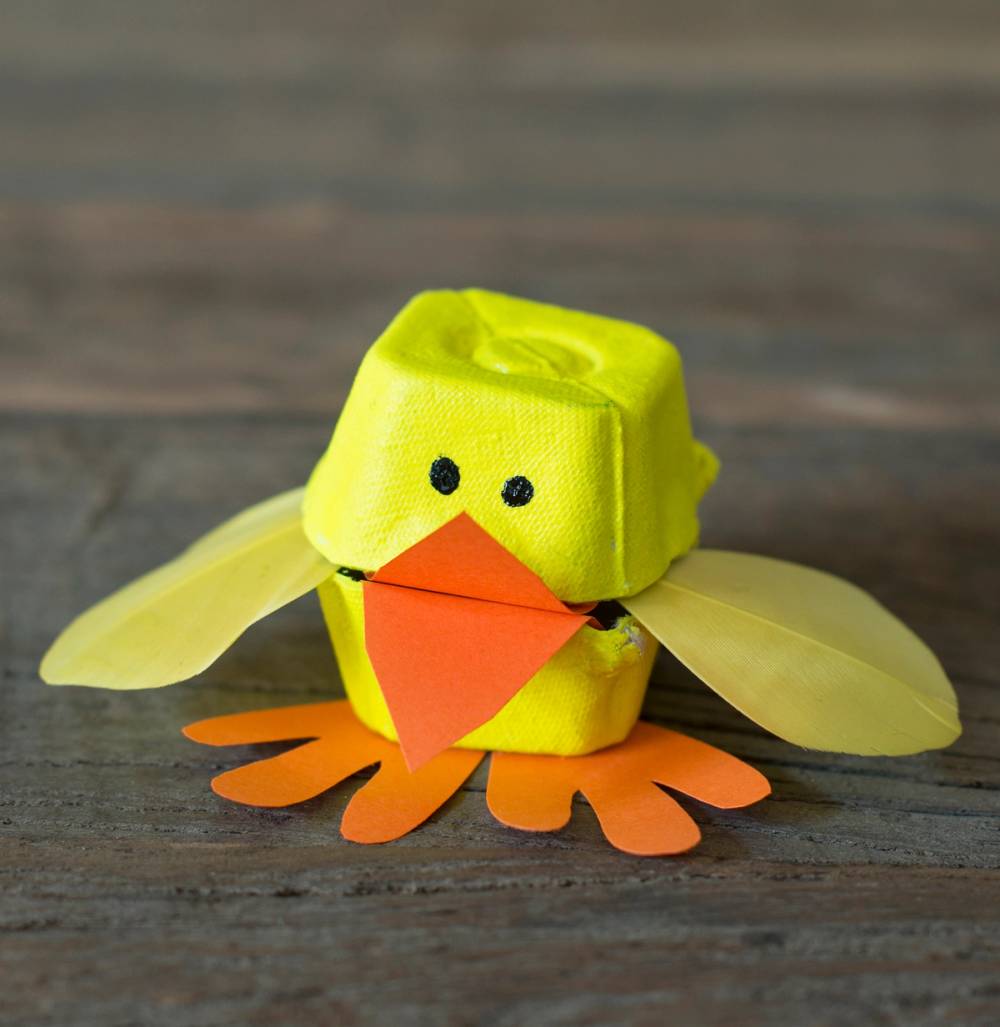 En kylling laget av en eggkartong med påskeaktige detaljer som vinger og føtter, plassert på en treoverflate.