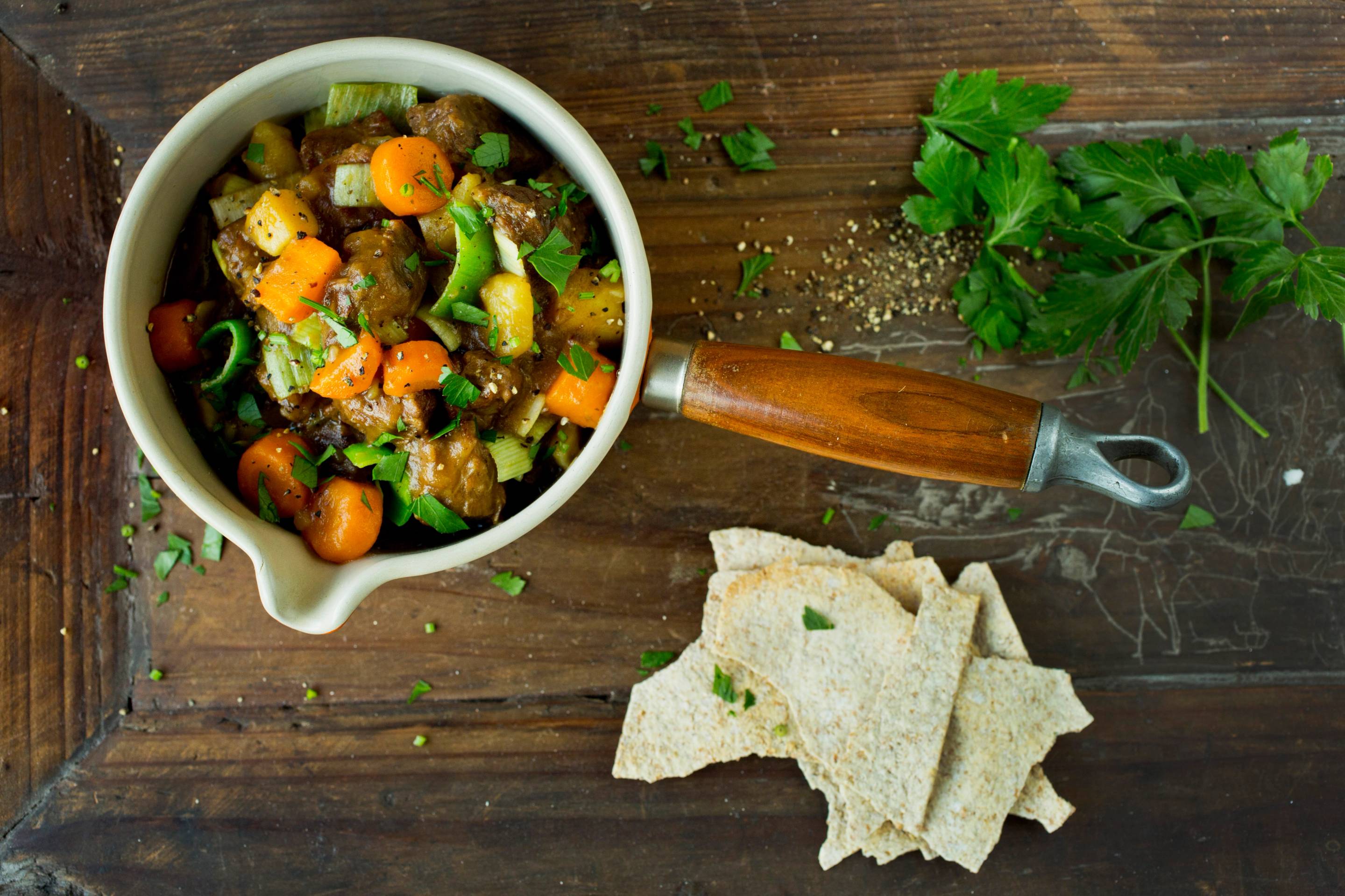 Lapskaus i en hvit bolle med gulrøtter, poteter og kjøtt, med flatbrød og frisk persille ved siden av på en treplate.