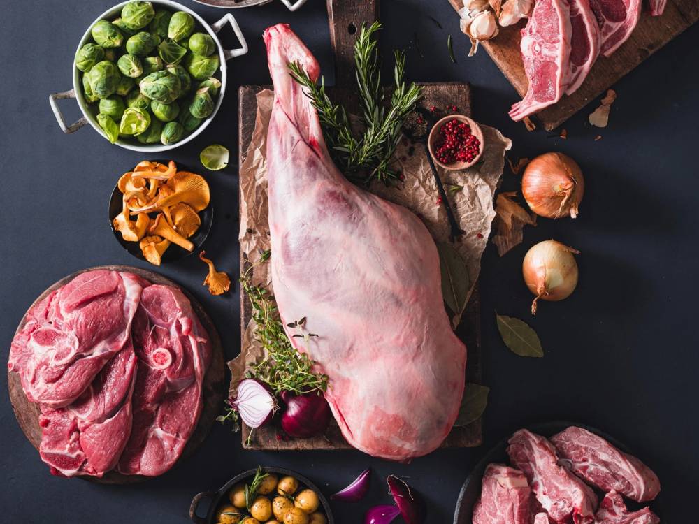 Ulike typer av kjøtt og grønnsaker på en mørk bakgrunn, inkludert lammebog, løk, hvitløk, og grønnsaker, klar til tilberedning.
