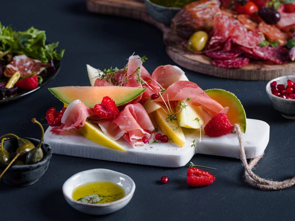 Spekeskinke og melon på en hvit serveringsplate med noen bær og oliven i bakgrunnen.