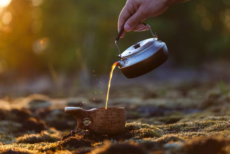 En person heller varm væske fra en kanne over et lite bål utendørs i skogen.