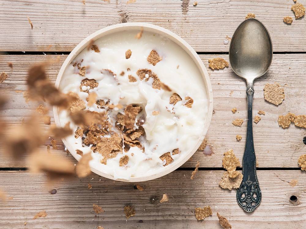 En skål med økologisk melk og kornbiter på et trebord, med en sølvskje ved siden av.