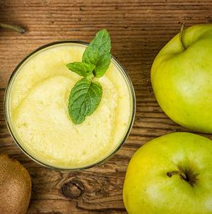 Et glass med frisk eple- og sitrusjuice med mynte på toppen, omgitt av grønne epler og en kiwi på et trebord.