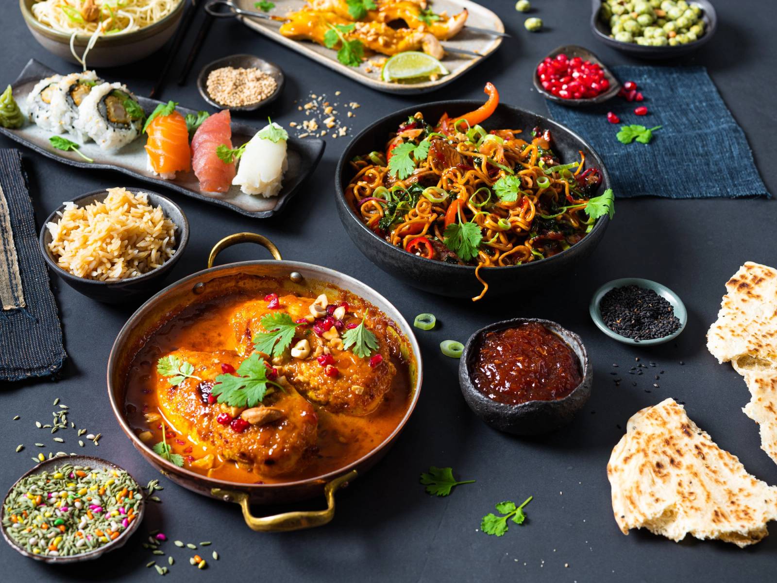 Et utvalg av asiatisk mat, inkludert curry, nudler og sushi, presentert på svarte tallerkener og skåler på et mørkt bord.