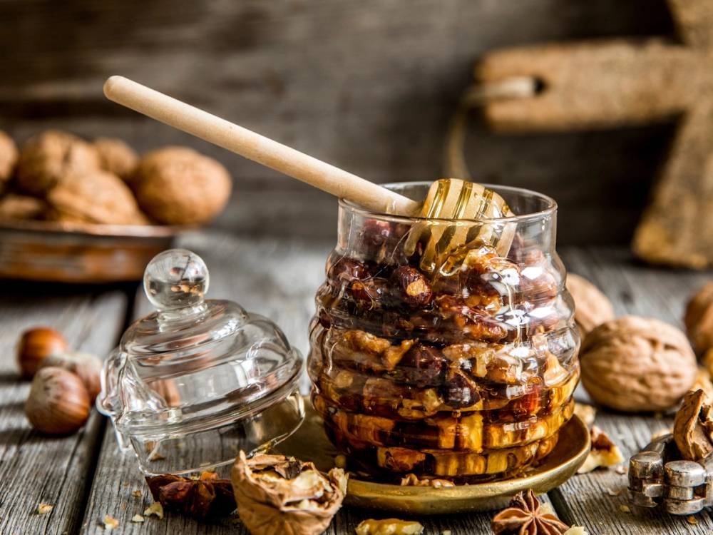 En glasskrukke med nøtter og honning, omgitt av flere nøtter på et bord.