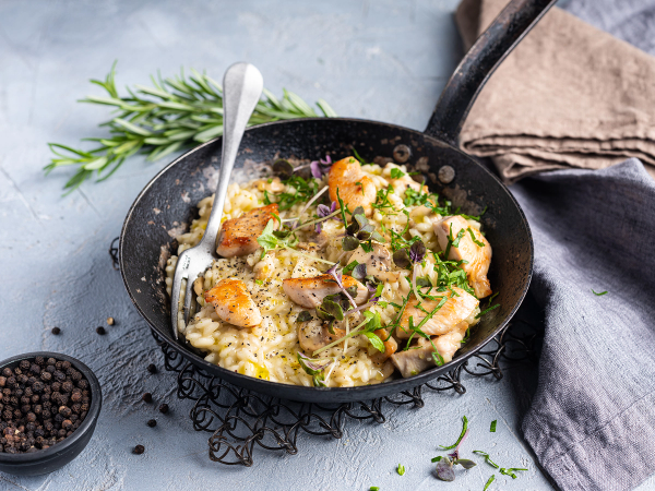 En italiensk kyllinggryte med ris i en svart stekepanne, pyntet med ferske urter og servert med en skje, på et lyst bord med en klut og en liten skål med pepper i bakgrunnen.