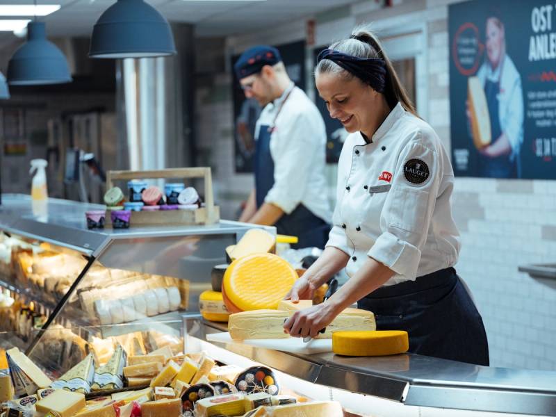 En person i hvit kokkeuniform skjærer ost ved en ostedisplay i en butikk eller på et marked, med andre personer i bakgrunnen.