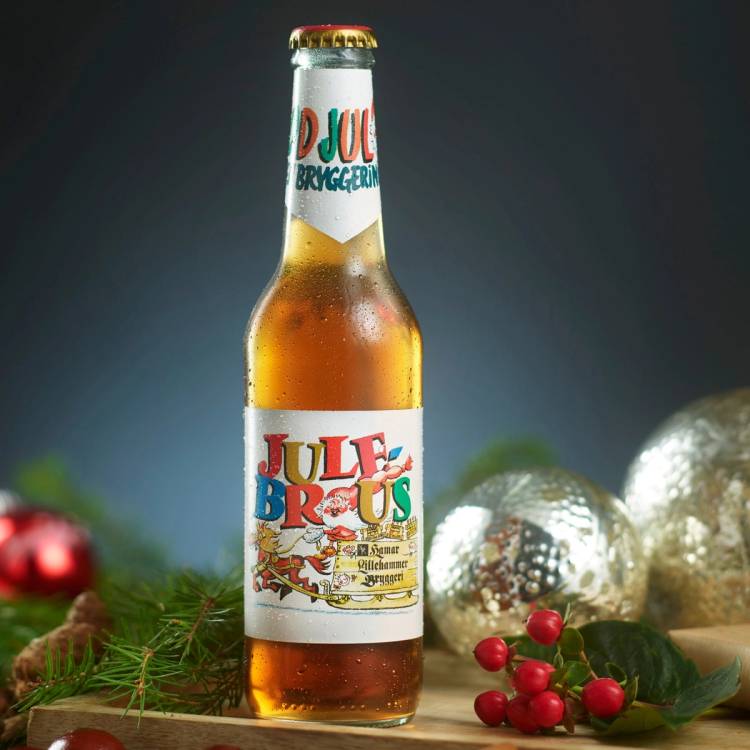 En flaske med julebrus står på et bord omgitt av juledekorasjoner som granbar, røde bær og juleballer.