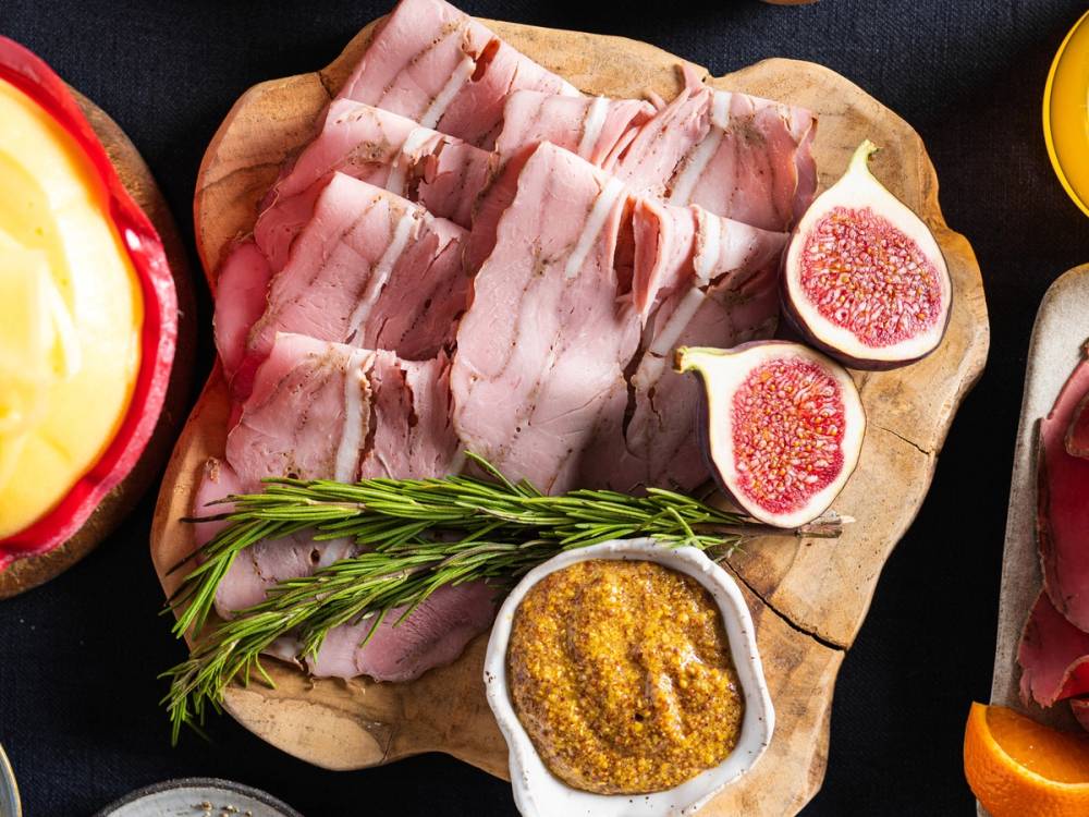 Spekemat med skiver av bacon, ferske fiken og en liten skål med sennep på et rundt trefat, omgitt av julefrokostrekvisita.