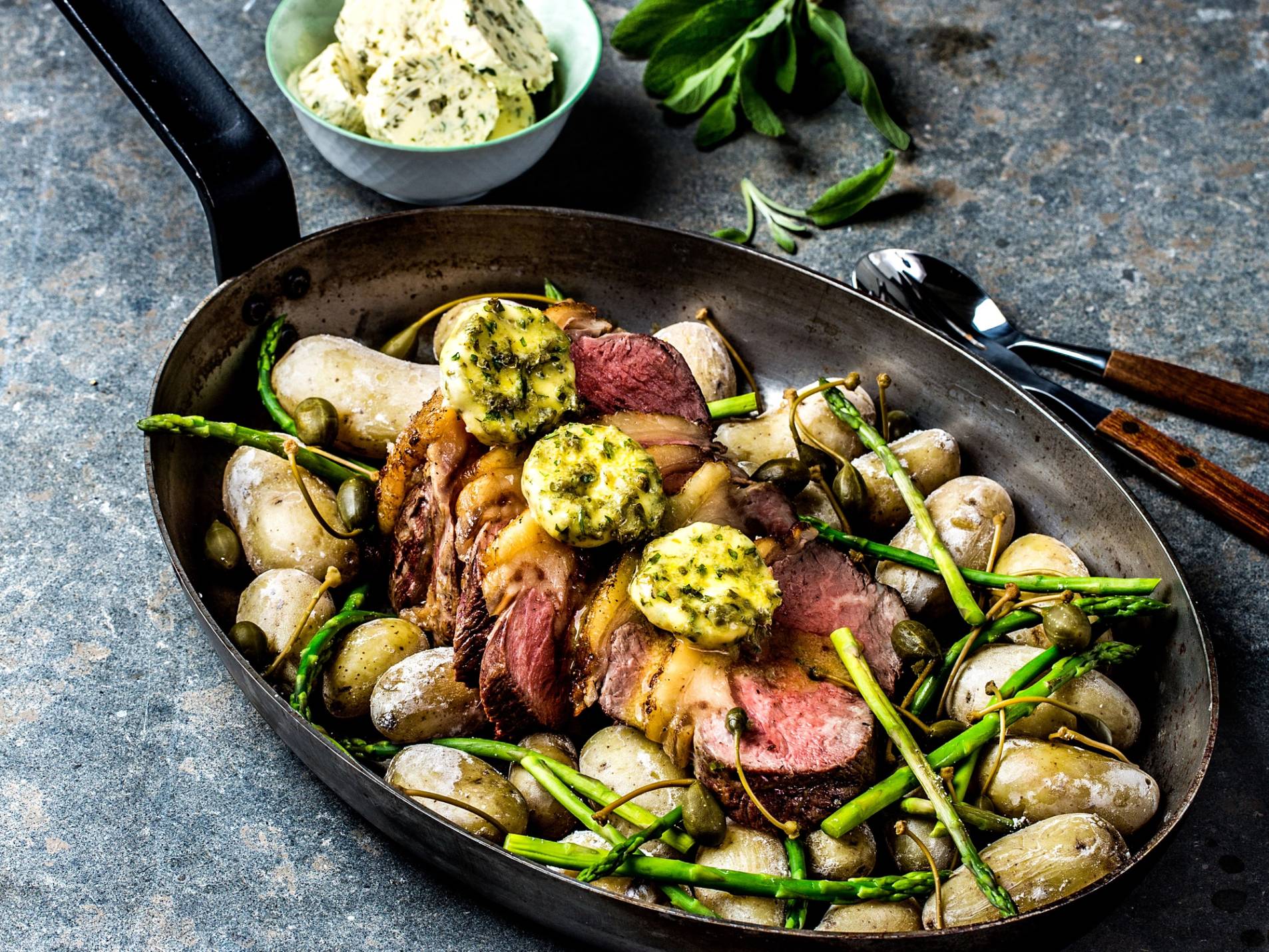 Helgrillet ytrefilet med fettlokk servert med grønne urter og små poteter på en svart stekepanne, med en skål med potetsalat i bakgrunnen.
