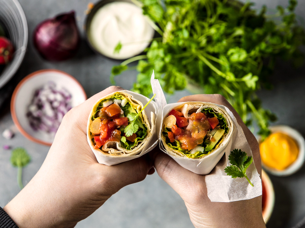 To tacorester som er rullet opp og fylt med kjøtt, salat og grønnsaker, holdt i hendene over en tallerken med mer salat og grønnsaker.