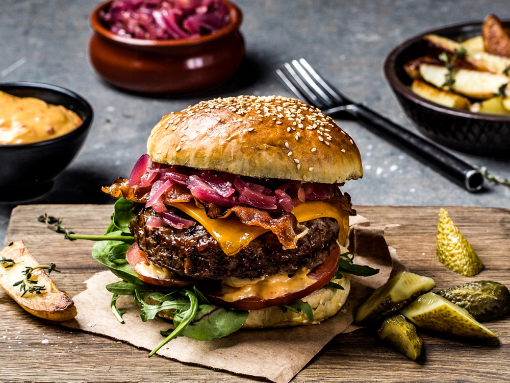 En saftig hamburger med cheddarost, bacon, salat og pickles på en treplate, med ulike sideretter rundt.