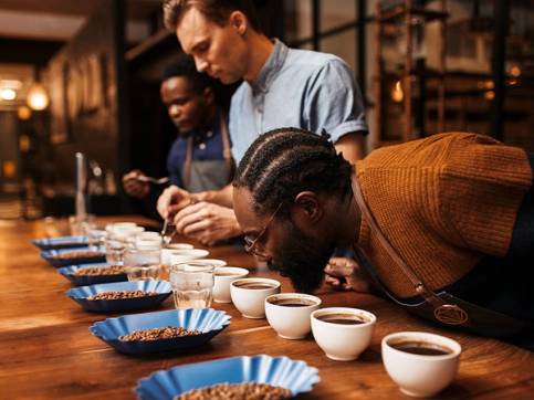 To personer som lukter på ulike typer kaffe i en kaffebar, med flere kopper og smaksprøver på bordet.