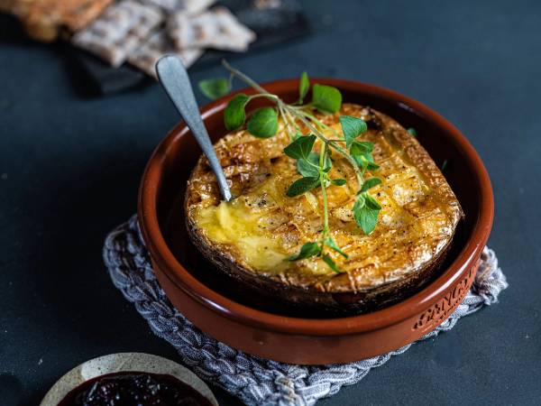 En gratinert ostefat med gyllen ost og grønt garnityr i en terrakottaskål, plassert på et svart underlag med en skje i.