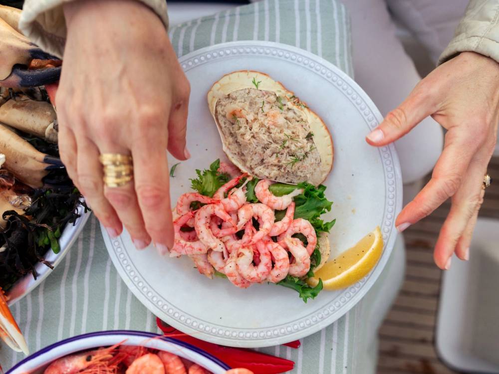 Hender som legger opp en porsjon reker med sitron på en tallerken med brød og grønnsaker, sett på et bord med en grønn og hvit stripet duk.