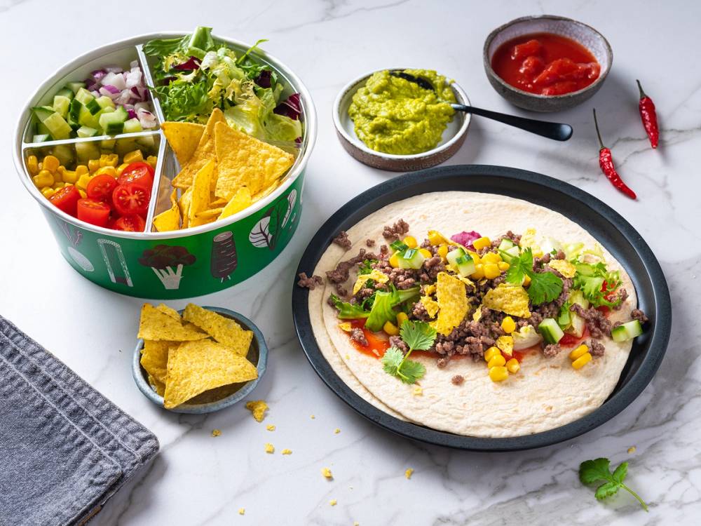 Tacos med kjøtt, mais, løk og koriander på en tallerken, salatbar med grønnsaker og chips ved siden av, samt guacamole og salsa i små skåler.