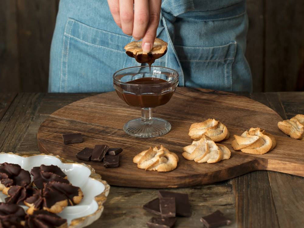 Person dypper en kransekaketopp i sjokolade mens de står ved et bord med flere andre kransekaketopper og sjokoladebiter.