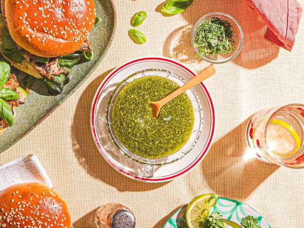 En bolle med grønn pesto og en skje oppi, omgitt av ulike matvarer og krydder på et bord.