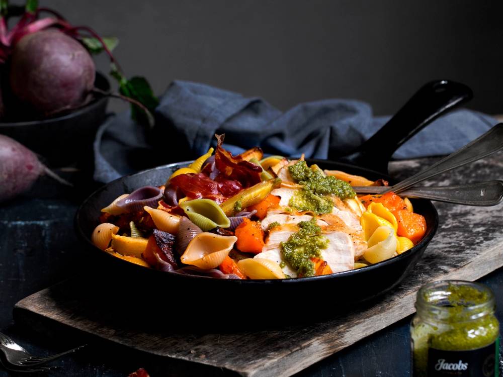 Pasta med kyllingbryst, bakt og servert i en svart stekepanne med grønnsaker og saus, på et treplate underlag.