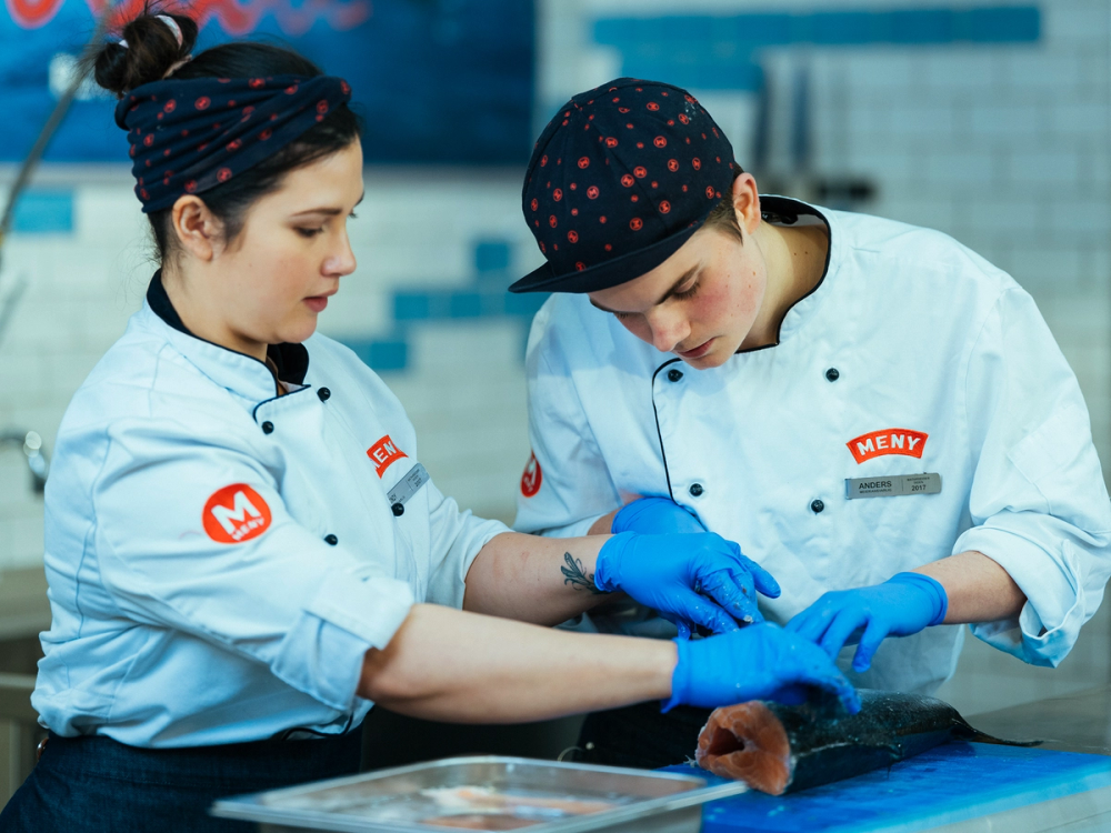 To ansatte i arbeidsklær og med blå hansker håndterer fisk i et kjøkkenmiljø.