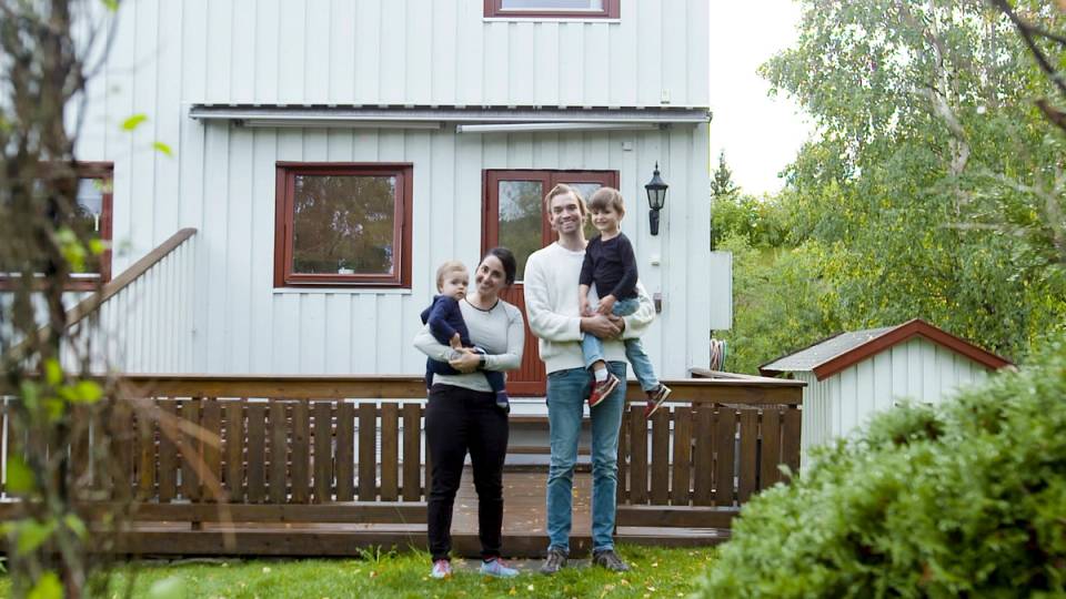 Fire personer står foran en hvit trehus med en liten terrasse, omgitt av grønt løv og trær.