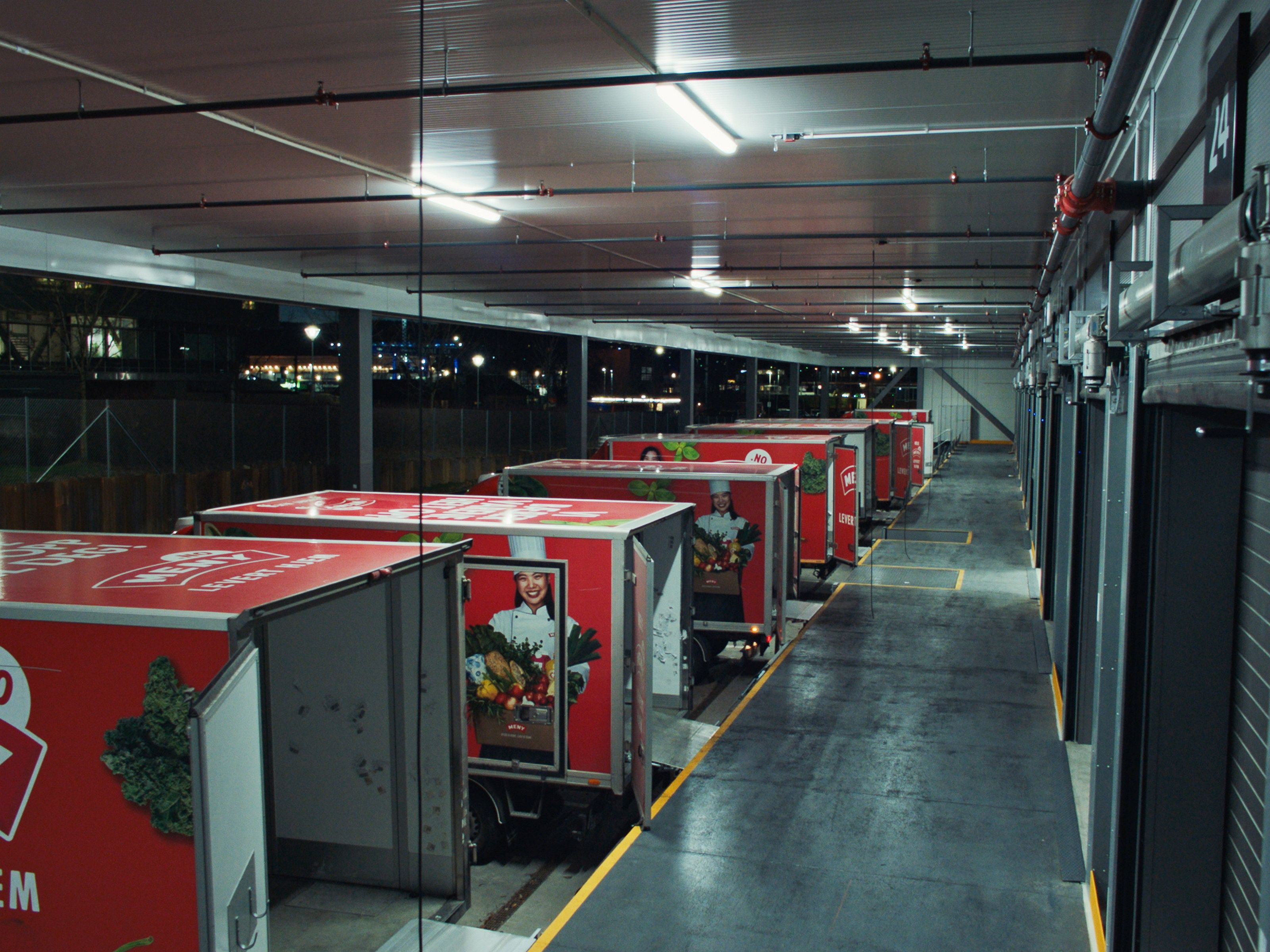 Leveringsbiler parkert i en innendørs parkeringsgarasje med grå gulv og tak, og røde biler med logoer på sidene.