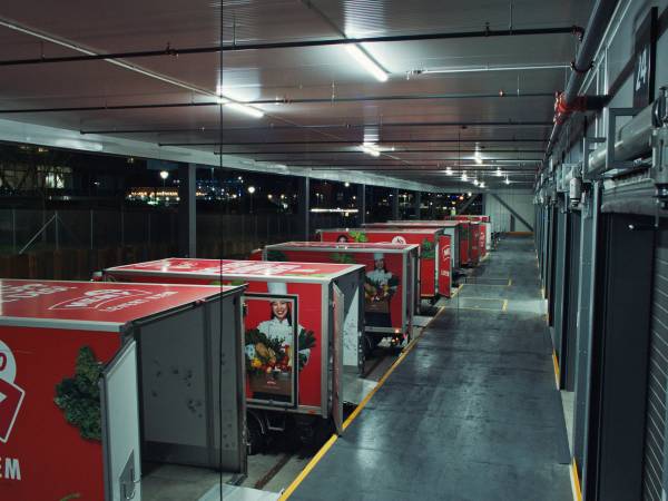 Leveringsbiler parkert i en innendørs parkeringsgarasje med grå gulv og tak, og røde biler med logoer på sidene.