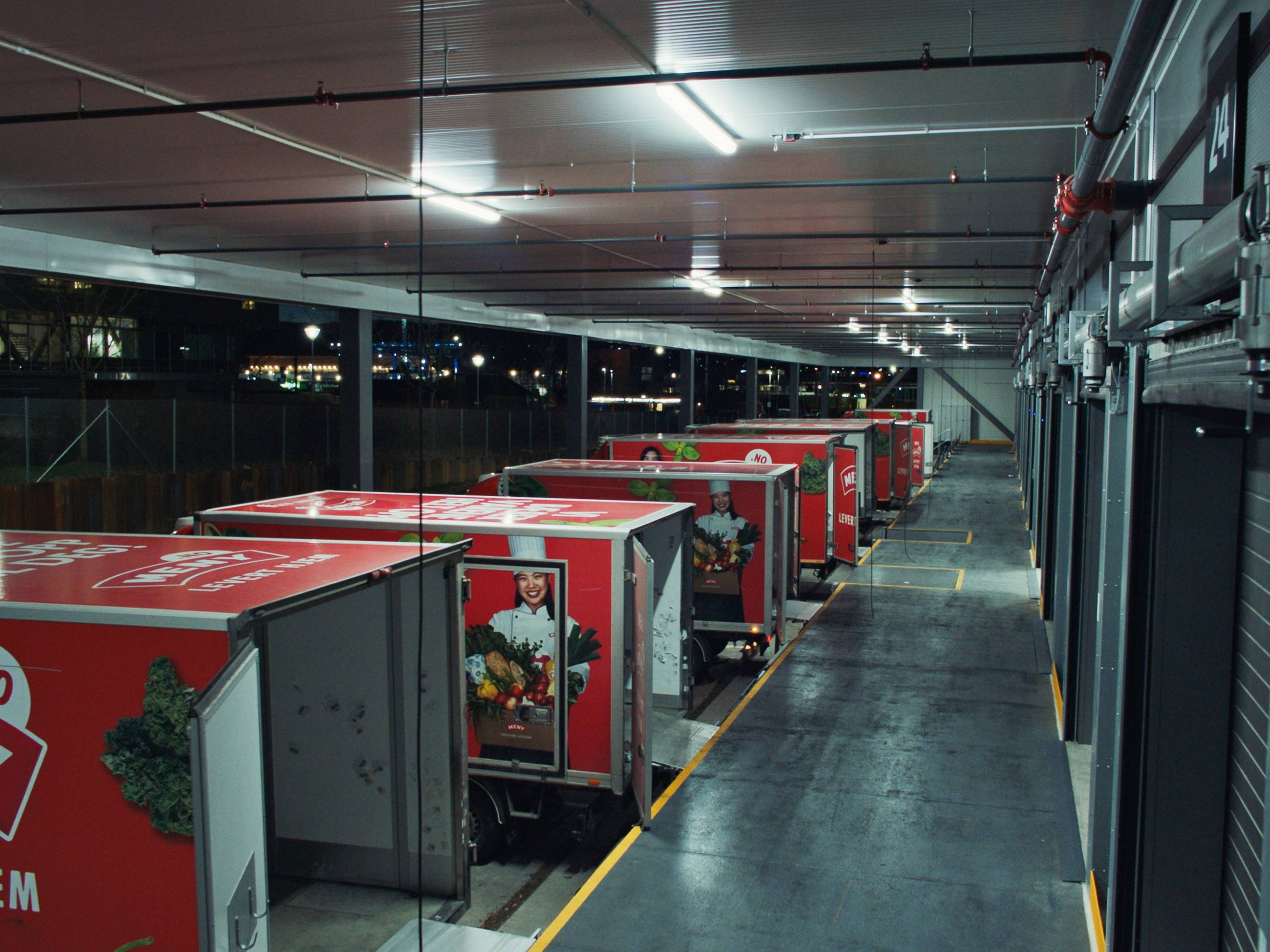 Leveringsbiler parkert i en innendørs parkeringsgarasje med grå gulv og tak, og røde biler med logoer på sidene.