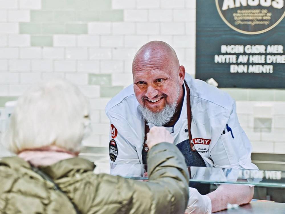 En person i hvit jakke snakker med en annen person på en kafé eller restaurant, med en vegg og en meny i bakgrunnen.