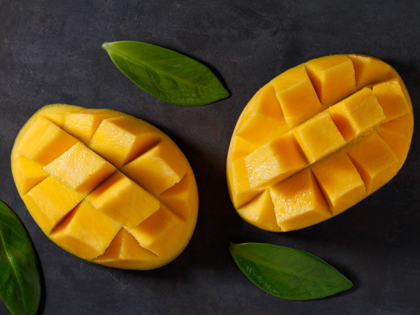 To halverte mangoer med terninger, omgitt av grønne blader på en mørk bakgrunn.
