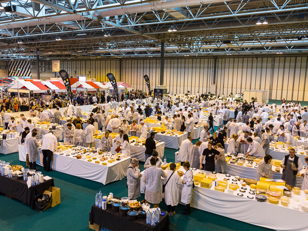 Stor matkonkurranse med mange deltakere i hvite kokkeuniformer som forbereder og presenterer ost på et stort innendørs arrangement.