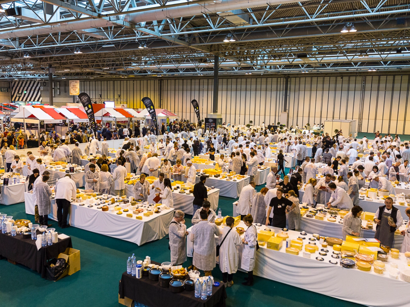 Stor matkonkurranse med mange deltakere i hvite kokkeuniformer som forbereder og presenterer ost på et stort innendørs arrangement.