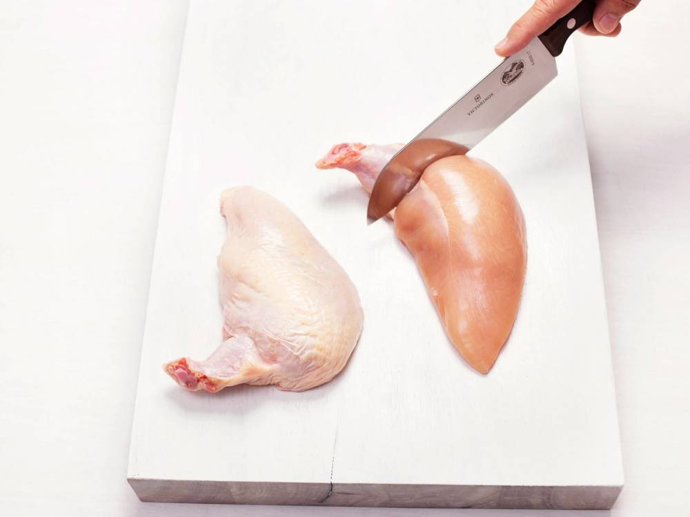 En person skjærer opp en rå kyllingbryst på et hvitt skjærebrett med en kniv.