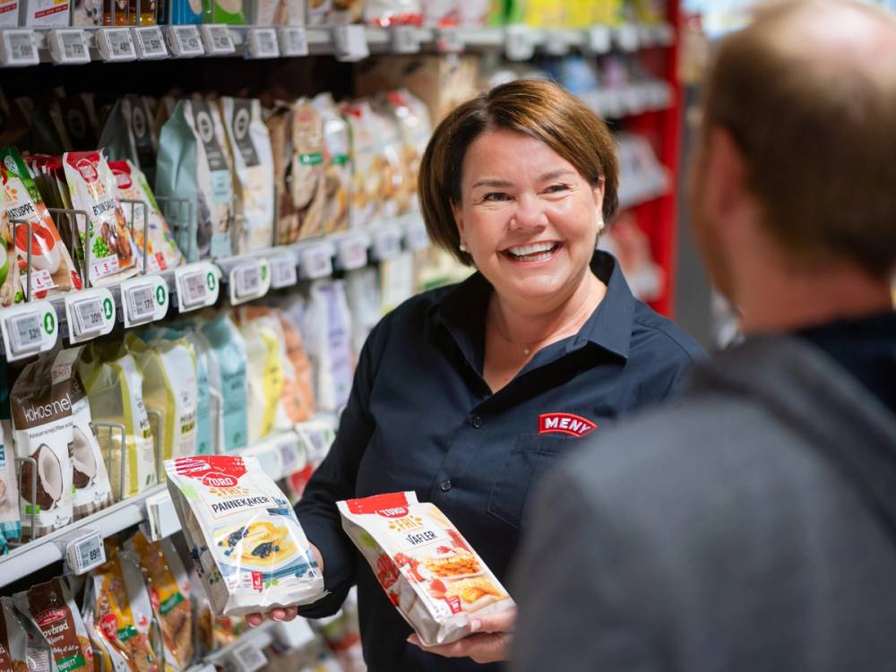 Sissel handler i en dagligvarebutikk med hyller fylt av ulike matvarer, og hun holder en pakke i hånden mens hun snakker med en mann.