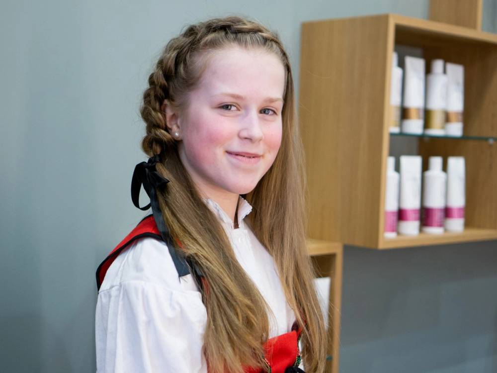 En jente med fletter og 17. mai-antrekk, står i et rom med hyller og produkter på bakgrunnen.