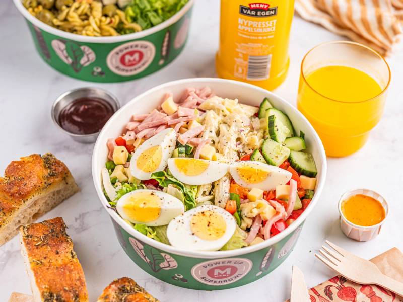 En salat med kokte egg, agurk, tomat og skinke i en grønn salatbar-kopp, omgitt av dressing, brød og en glass med appelsinjuice.