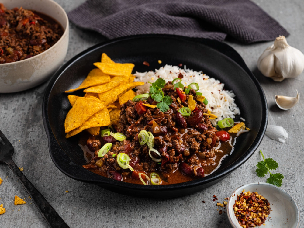 Chili con carne servert med ris, tortilla chips og fersk koriander på en svart tallerken.
