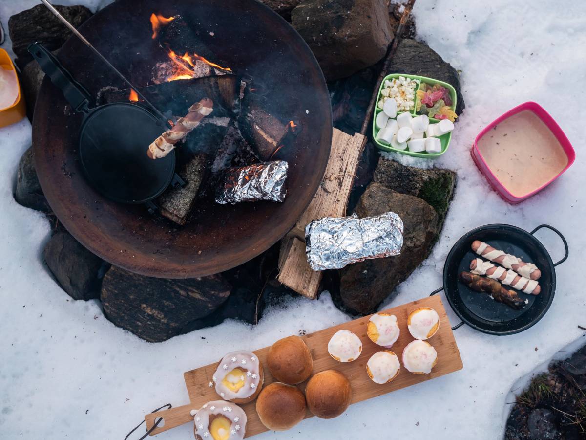 Sissel Fri For-14.jpg viser en utendørs grilling med mat som pølser, poteter og grønnsaker på et bord i snøen.