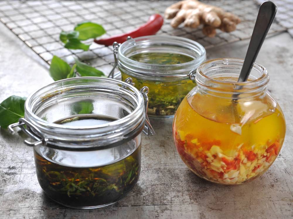 Tre glass med ulike marinader, en med olje og urter, en med en klar væske og en med en gul væske med krydder, på et trebenk med urter og chili i bakgrunnen.