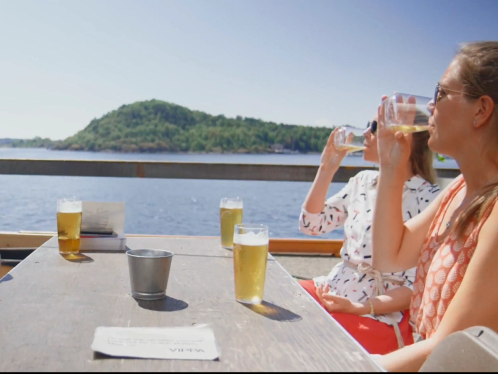 To personer nyter drikke ved et utendørsbord med utsikt over vann og en øy i bakgrunnen.